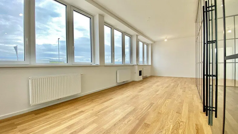 Heller, leerer Büroraum mit großen Fenstern, Holzfußboden und modernen Glaswänden, die viel Tageslicht hereinlassen.