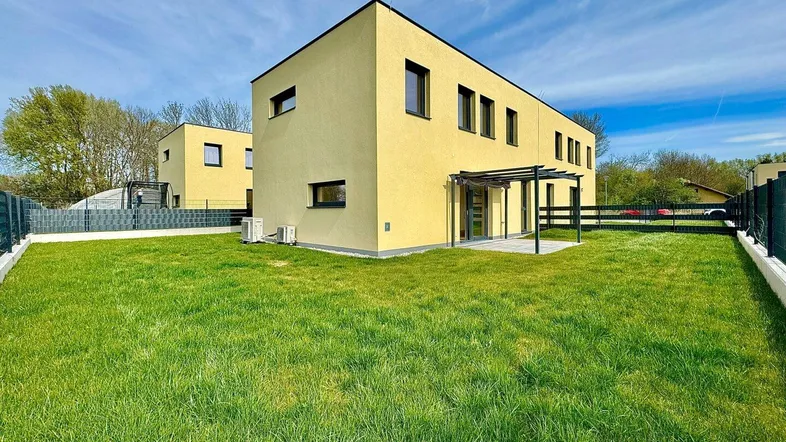 Modernes Reihenhaus mit gelber Fassade, großem Rasen und Pergola-Terrasse unter blauem Himmel.