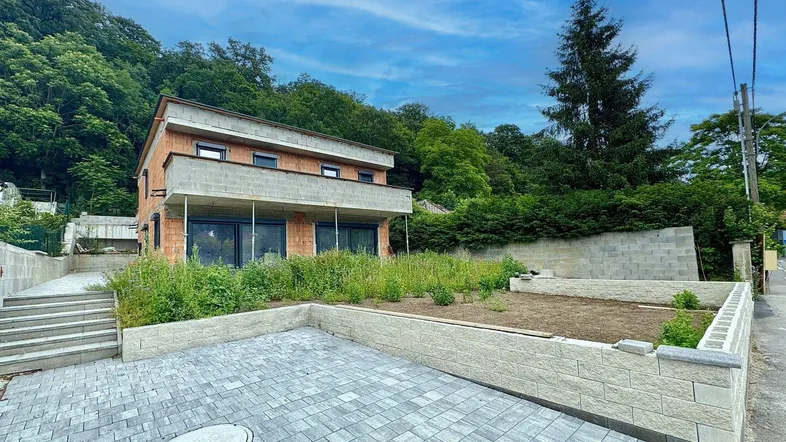 Neubauhaus in Hanglage mit gepflastertem Vorplatz und vorbereiteten Gartenbeeten, umgeben von viel Grün.