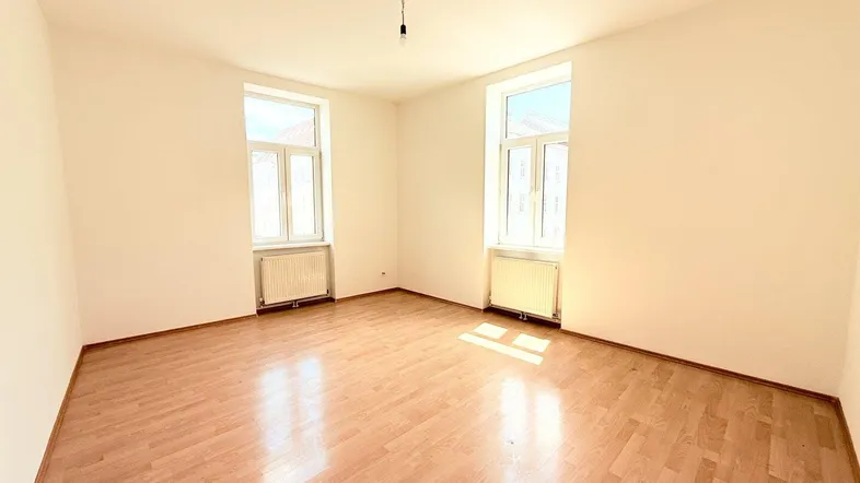 Heller, leerer Raum mit Laminatboden, zwei großen Fenstern und Heizkörpern unter den Fenstern.