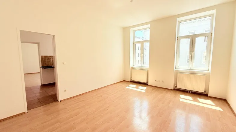 Helles, leeres Zimmer mit Laminatboden und zwei Fenstern, die viel Tageslicht hereinlassen.