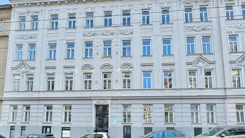 Klassische Fassade eines Mehrfamilienhauses mit mehreren Stockwerken und geparkten Autos davor.