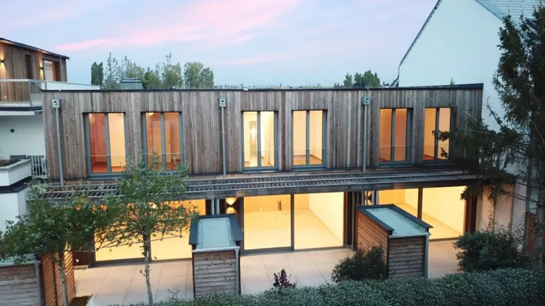Moderne Holzfassade eines Gebäudes mit großen Fenstern und beleuchteter Terrasse am Abend.