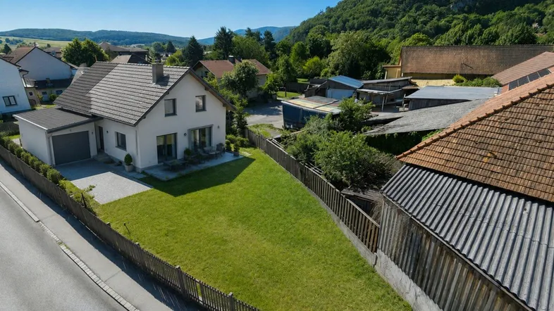 Luftaufnahme eines Einfamilienhauses mit gepflegtem Garten, Garage und Blick auf die umliegende Hügellandschaft.