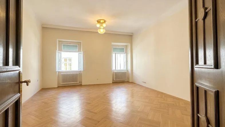 Helles, leeres Zimmer mit Fischgrätparkett, hohen Decken und zwei Fenstern.