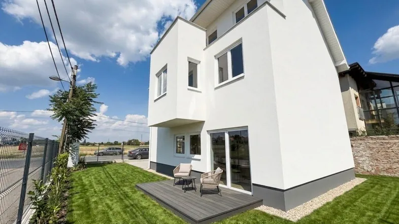Modernes Einfamilienhaus mit weißer Fassade, kleiner Holzterrasse und gepflegtem Rasen unter blauem Himmel.