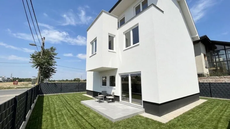 Moderne Hausfassade mit weißem Putz, kleiner Terrasse und gepflegtem Rasen unter blauem Himmel.