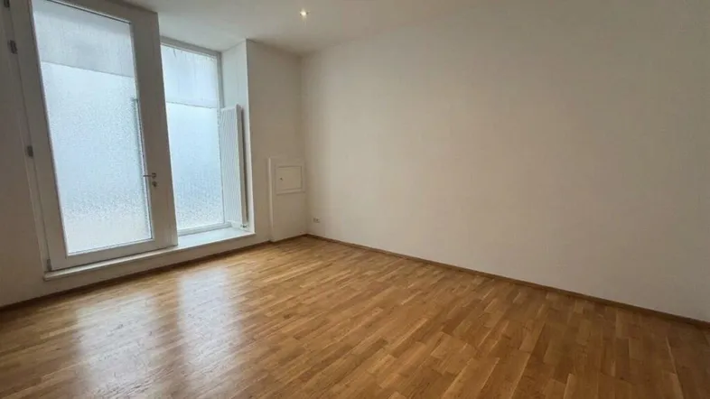 Heller, leerer Raum mit warmem Holzboden, weißen Wänden und großen Fenstern für viel Tageslicht.