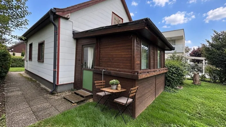 Hausfassade mit weißer Verkleidung, hölzernem Wintergarten und einem gepflegten Garten mit Sitzgelegenheit.