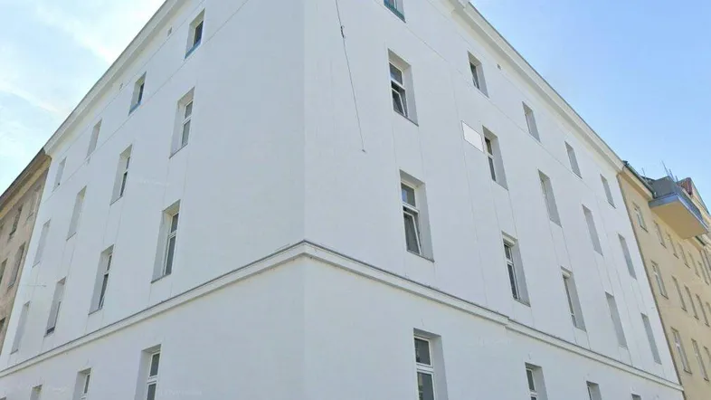 Weißes Mehrfamilienhaus mit vielen Fenstern unter blauem Himmel, an einer Straße gelegen.