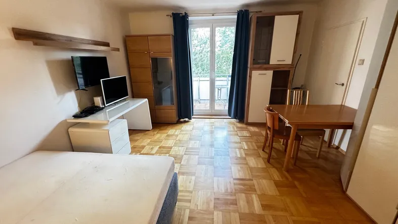 Helles Studio-Apartment mit Parkettboden, Schlafbereich, Schreibtisch und Zugang zum Balkon.