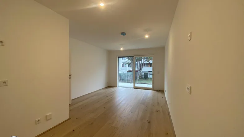 Helles, leeres Wohnzimmer mit Parkettboden und Zugang zu einem Balkon oder einer Terrasse.