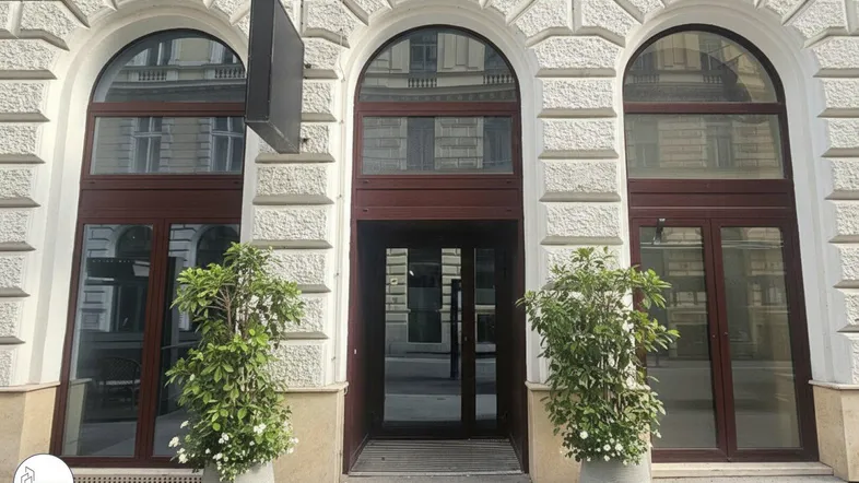 Eingangsbereich eines Gebäudes mit klassischer Fassade, großen Rundbogenfenstern und zwei Pflanzen.