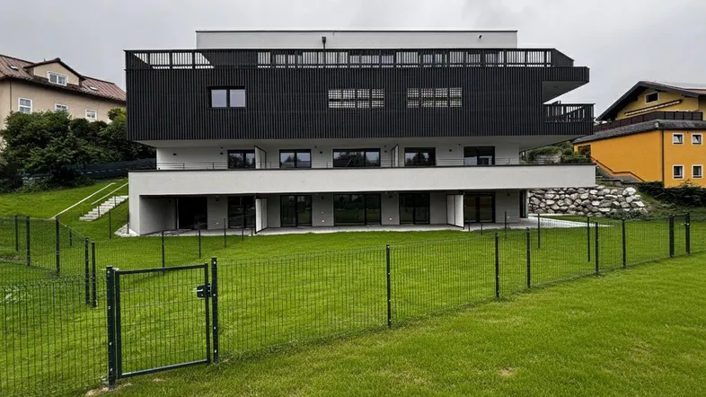 Moderne Wohnanlage mit schwarzer Lamellenfassade und weißem Sockelgeschoss, umgeben von einer Grünfläche.