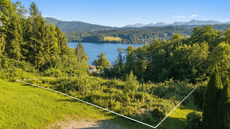 Panoramablick auf ein grünes Grundstück mit See und Bergen im Hintergrund, umrandet von Bäumen.
