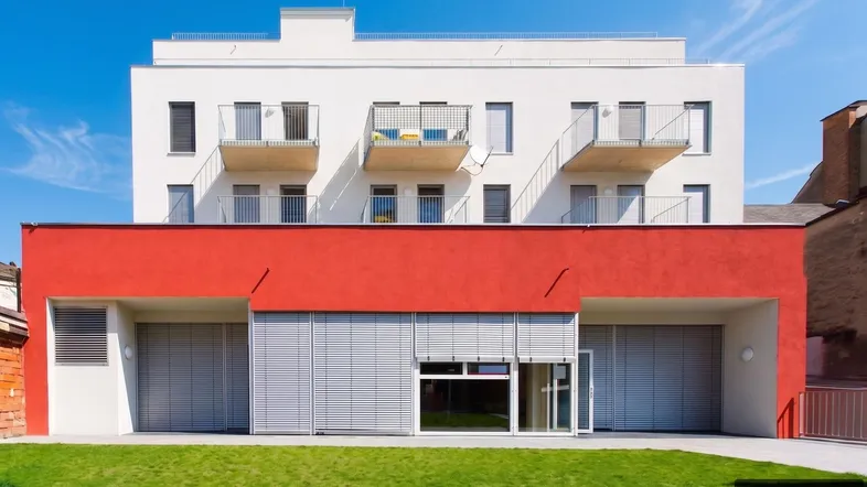 Modernes Wohngebäude mit weißer Fassade, roten Akzenten und kleinen Balkonen über einer Grünfläche.