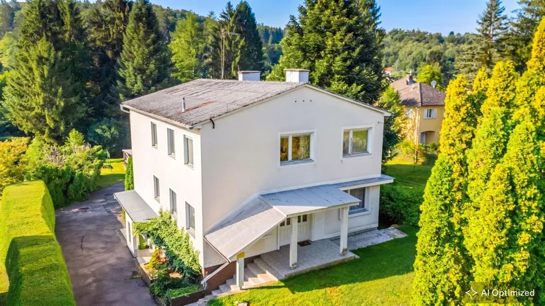 Weißes Einfamilienhaus mit grauem Dach, umgeben von üppigem Grün und hohen Bäumen.