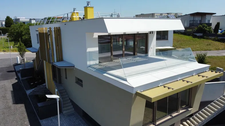 Moderne Architektur mit großer Terrasse, Glasgeländer und viel Tageslicht unter blauem Himmel.