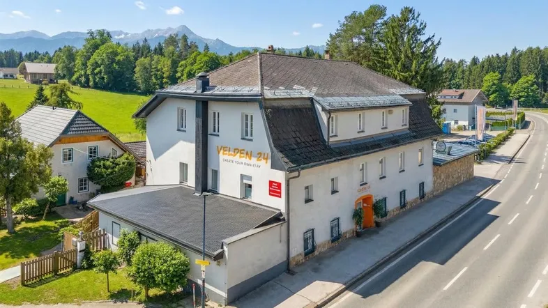 Gebäudeansicht von der Straße mit grüner Umgebung und majestätischen Bergen im Hintergrund.