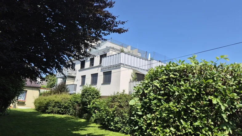 Mehrfamilienhaus mit moderner Architektur, umgeben von Grünflächen und Bäumen unter blauem Himmel.