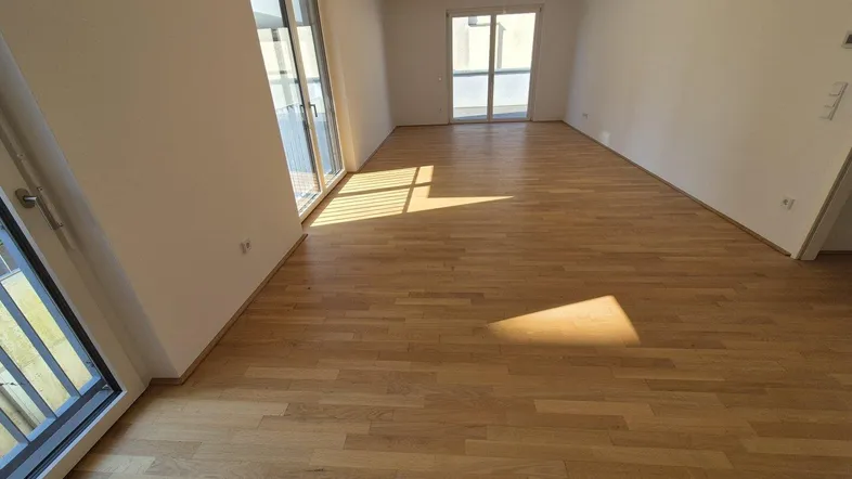 Helles, leeres Zimmer mit Holzboden und großen Fenstern, die viel Tageslicht hereinlassen.