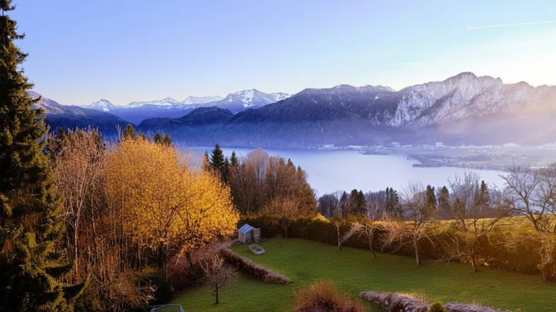 Herbstliche Gartenlandschaft mit Pool und See, umrahmt von bunten Bäumen und schneebedeckten Berggipfeln.