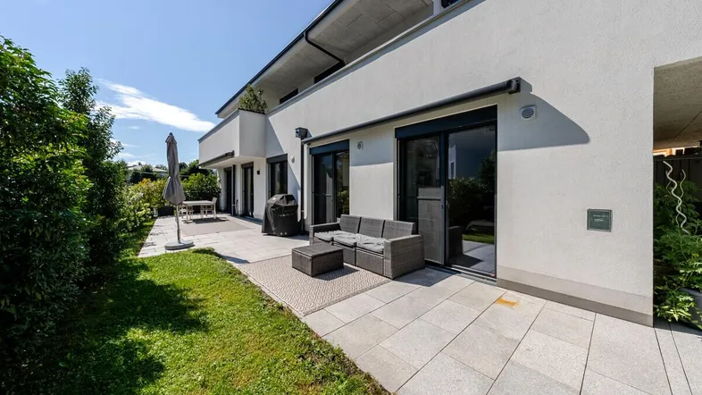 Moderne Terrasse mit Sitzgelegenheiten und Rasenfläche, umgeben von grüner Bepflanzung an einem sonnigen Tag.