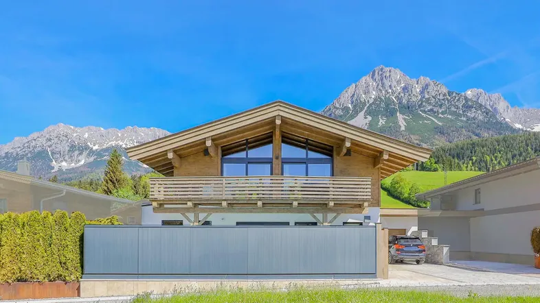 Modernes Holzhaus mit Balkon und beeindruckendem Bergblick unter blauem Himmel.