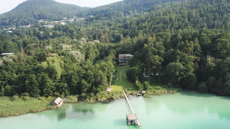 Luftaufnahme eines Hauses am See, umgeben von Wald und Bergen, mit eigenem Bootssteg.