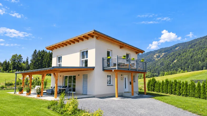 Modernes Einfamilienhaus mit überdachter Terrasse und Balkon in ländlicher Umgebung mit Bergblick unter blauem Himmel.