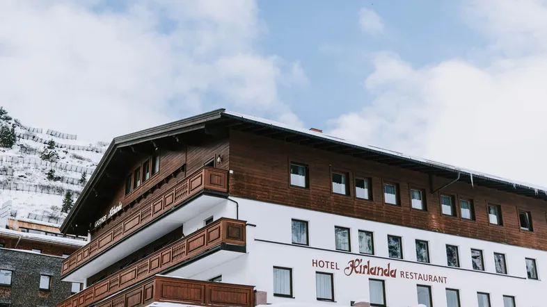 Ansicht des Hotels Kirlana mit traditioneller Holzfassade und schneebedeckten Bergen im Hintergrund.