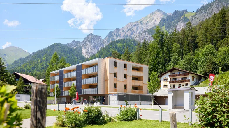 Modernes Gebäude mit Holzfassade und Balkonen, umgeben von grünen Bergen und Bäumen.