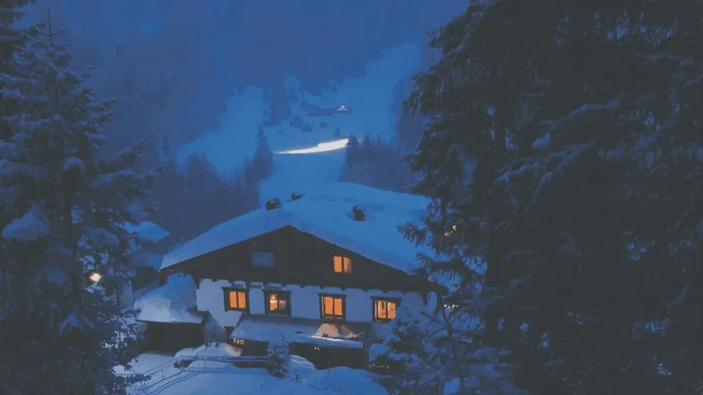 Traditionelles Chalet im Schnee bei Nacht, umgeben von verschneiten Bäumen und Bergen.