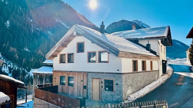 Modernes Haus in den Bergen mit Holz- und Steinfassade, umgeben von Schnee und Sonnenschein.