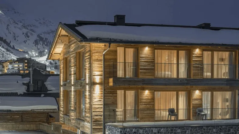 Modernes Holzchalet mit beleuchteten Balkonen und verschneiter Bergkulisse bei Nacht.