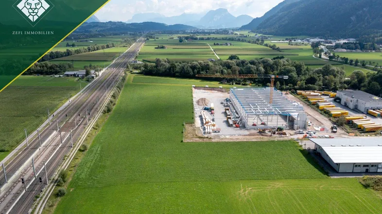 Luftaufnahme einer Baustelle für ein großes Industriegebäude, umgeben von grünen Feldern und Bergen.