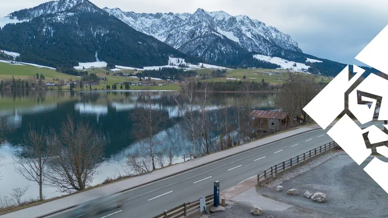 Panoramablick auf einen See mit schneebedeckten Bergen im Hintergrund und einer Straße im Vordergrund.