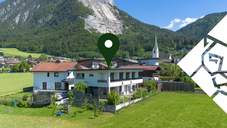 Modernes Haus mit Garten und Spielplatz vor einer beeindruckenden Bergkulisse und Dorfkirche.
