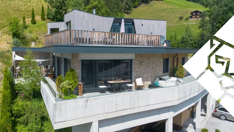 Modernes Gebäude am Hang mit mehreren Terrassen, Holzverkleidung und Steinmauerwerk, umgeben von grüner Natur.