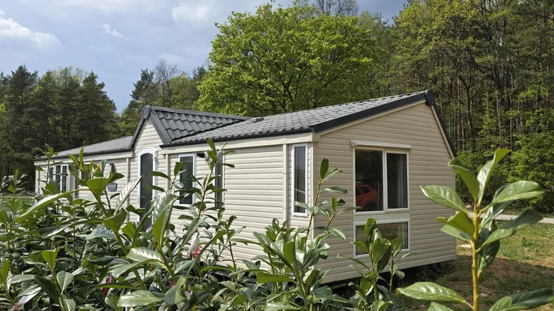 Modernes Mobilheim mit beiger Fassade und dunklem Dach, umgeben von grüner Natur und Bäumen.
