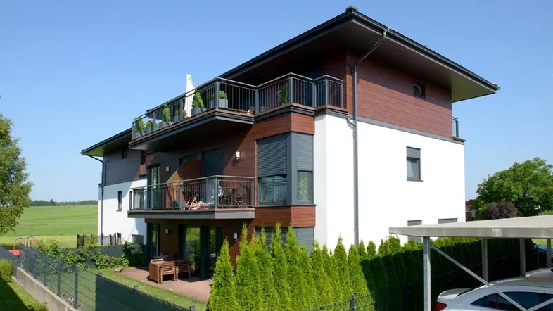 Modernes Mehrfamilienhaus mit weißer Fassade, Holzverkleidung, Balkonen und Terrasse, umgeben von Grünflächen.