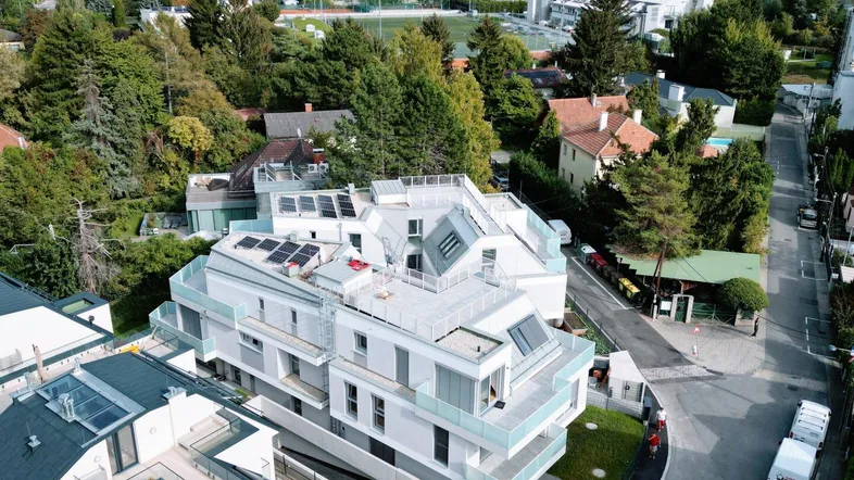 Moderne Wohnanlage mit Flachdach und Solarpaneelen, umgeben von viel Grün und einem See im Hintergrund.