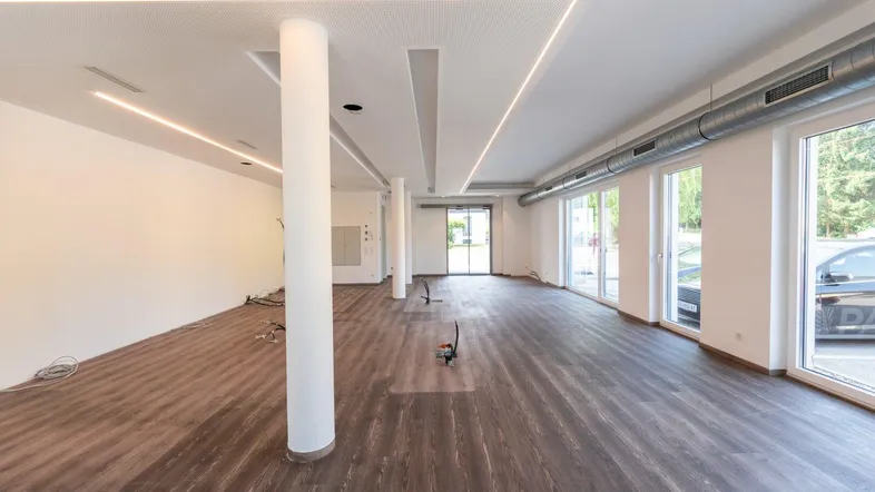 Geräumiger, leerer Gewerberaum mit großen Fenstern, modernen Deckenleuchten und dunklem Laminatboden, bereit für den Ausbau.