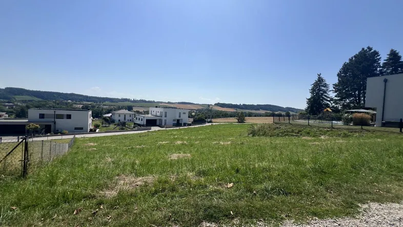 Blick über ein sonniges Wiesengrundstück mit modernen Häusern und hügeliger Landschaft im Hintergrund.