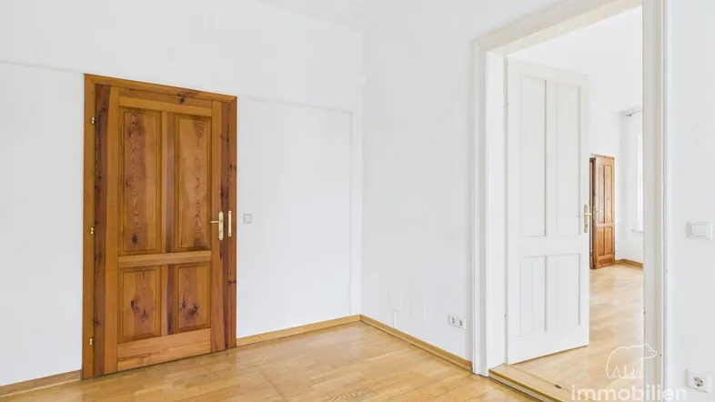Helles Zimmer mit Holzboden und zwei Holztüren, eine davon offen, die andere geschlossen.