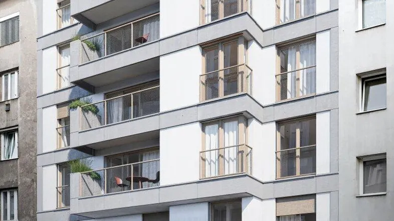 Moderne Wohnhausfassade mit gestaffelten Balkonen und großen Fenstern, umgeben von urbaner Architektur.
