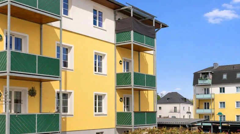 Gelbe Fassade eines Mehrfamilienhauses mit grünen Balkonen unter blauem Himmel.