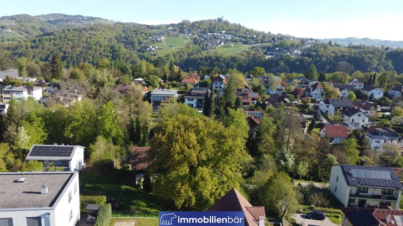 Luftaufnahme einer Wohnsiedlung mit viel Grün und bewaldeten Hügeln im Hintergrund.