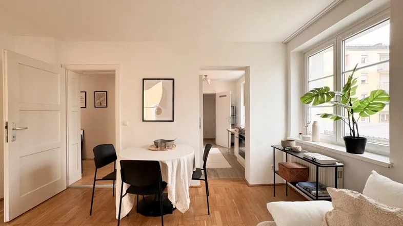 Heller Wohn- und Essbereich mit Holzboden, großem Fenster und Zugang zu weiteren Räumen.