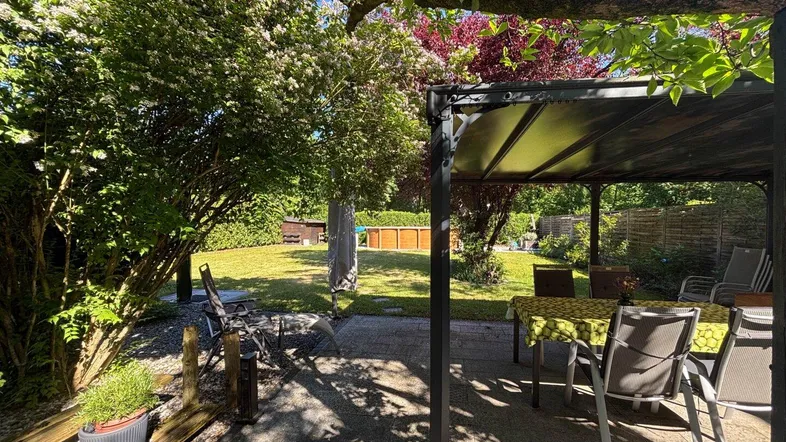 Sonniger Garten mit überdachtem Sitzbereich, Rasenfläche und einem Aufstellpool im Hintergrund.
